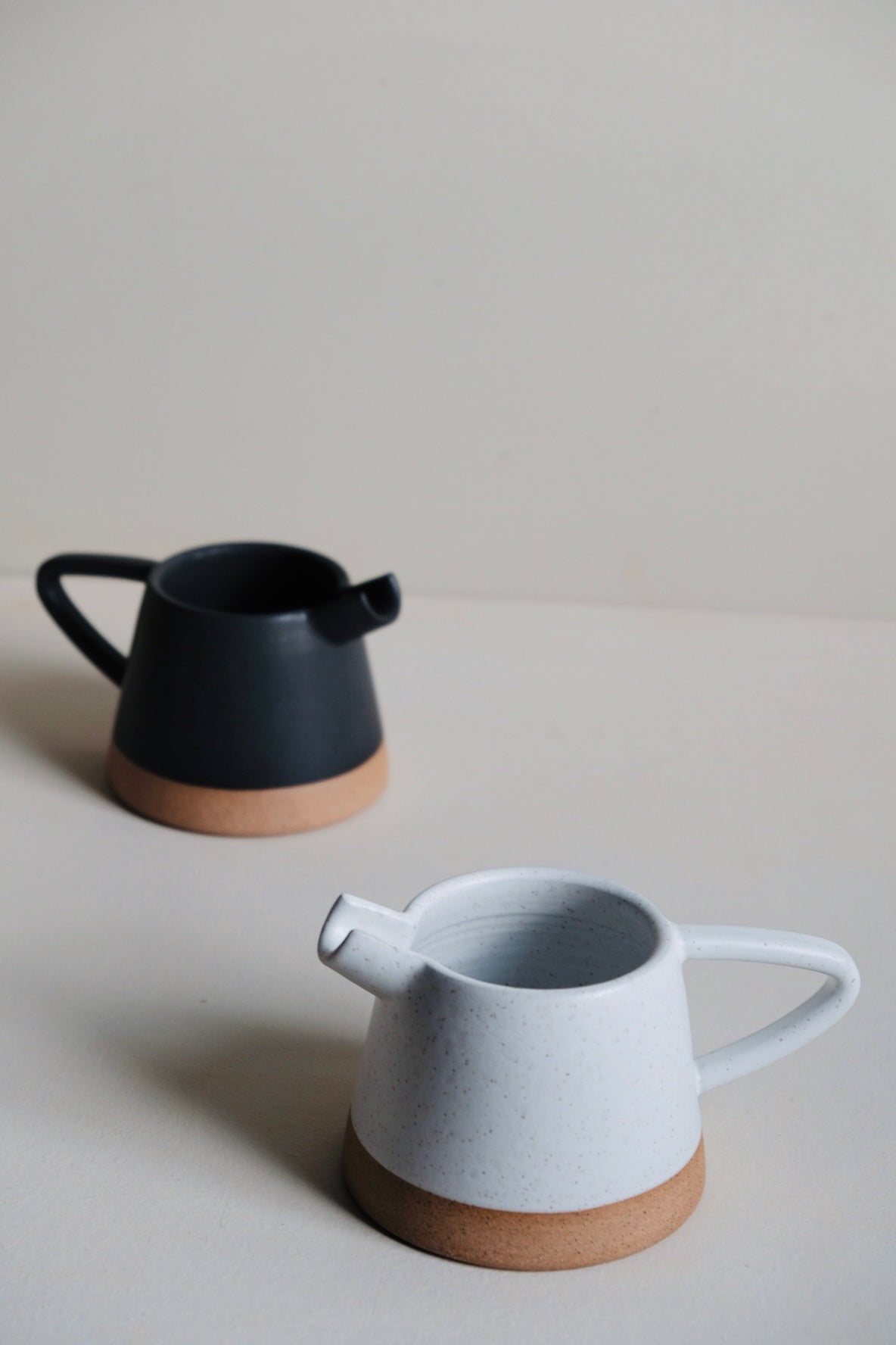 Two ceramic pitchers, one black and one white with brown base, on a light gray surface.